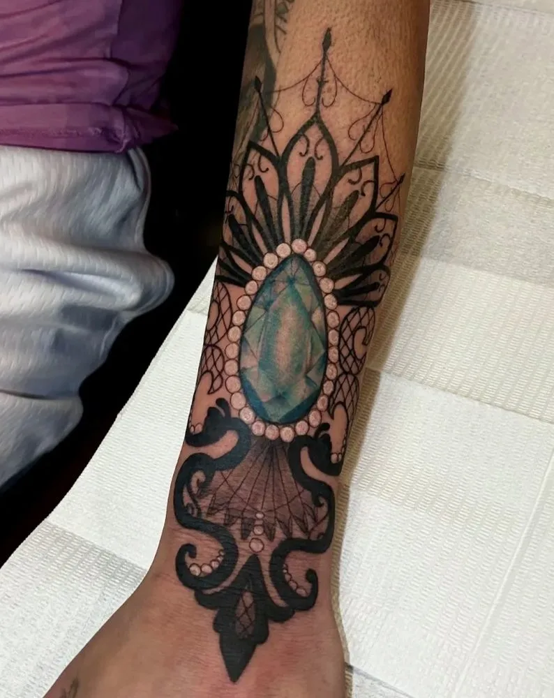 Black-and-grey ornamental lacework tattoo framing a teal gemstone
