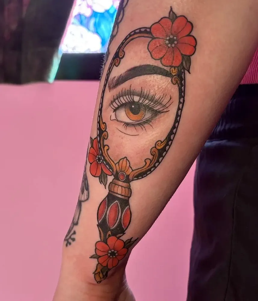 Traditional hand mirror tattoo reflecting an eye, framed with red flowers
