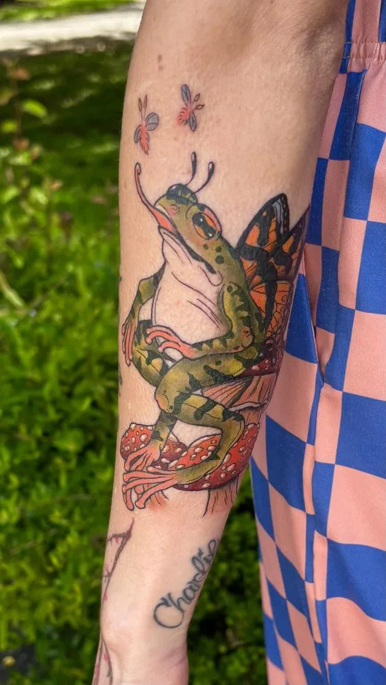 Neo-traditional frog with monarch butterfly wings perched on red spotted mushrooms tattoo