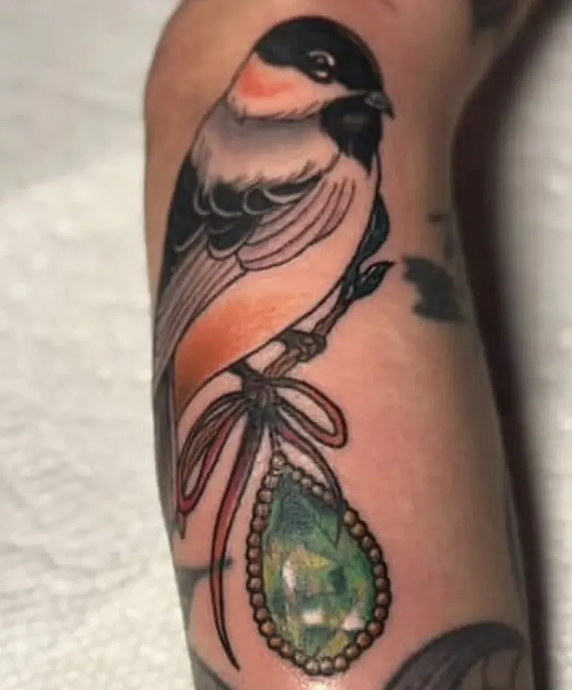 Songbird perched on a ribbon holding a green crystal pendant tattoo